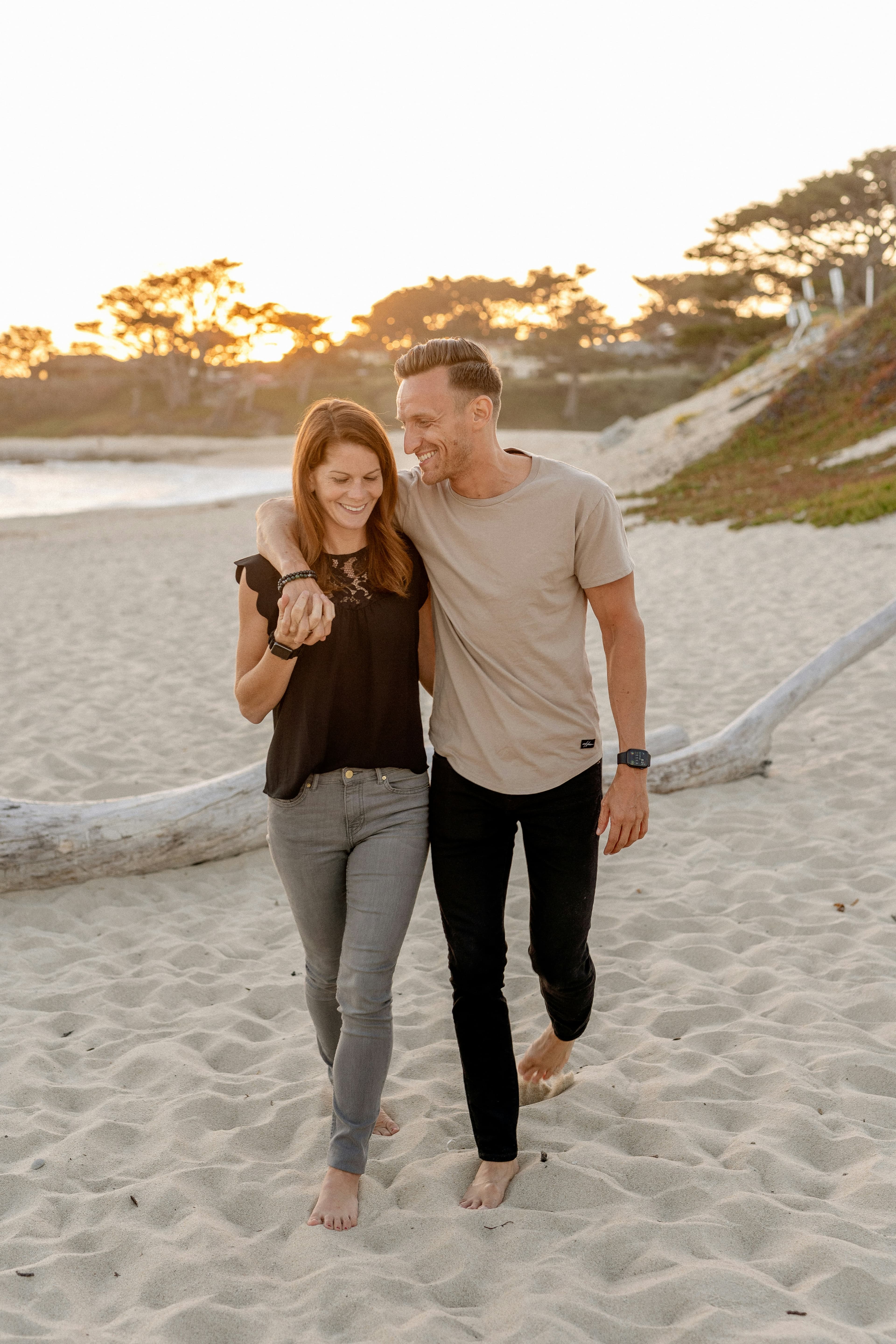 A couple walking barefoot on a sandy beach at sunset, laughing together. She wears a black top and gray jeans, he wears a beige t-shirt and black pants.