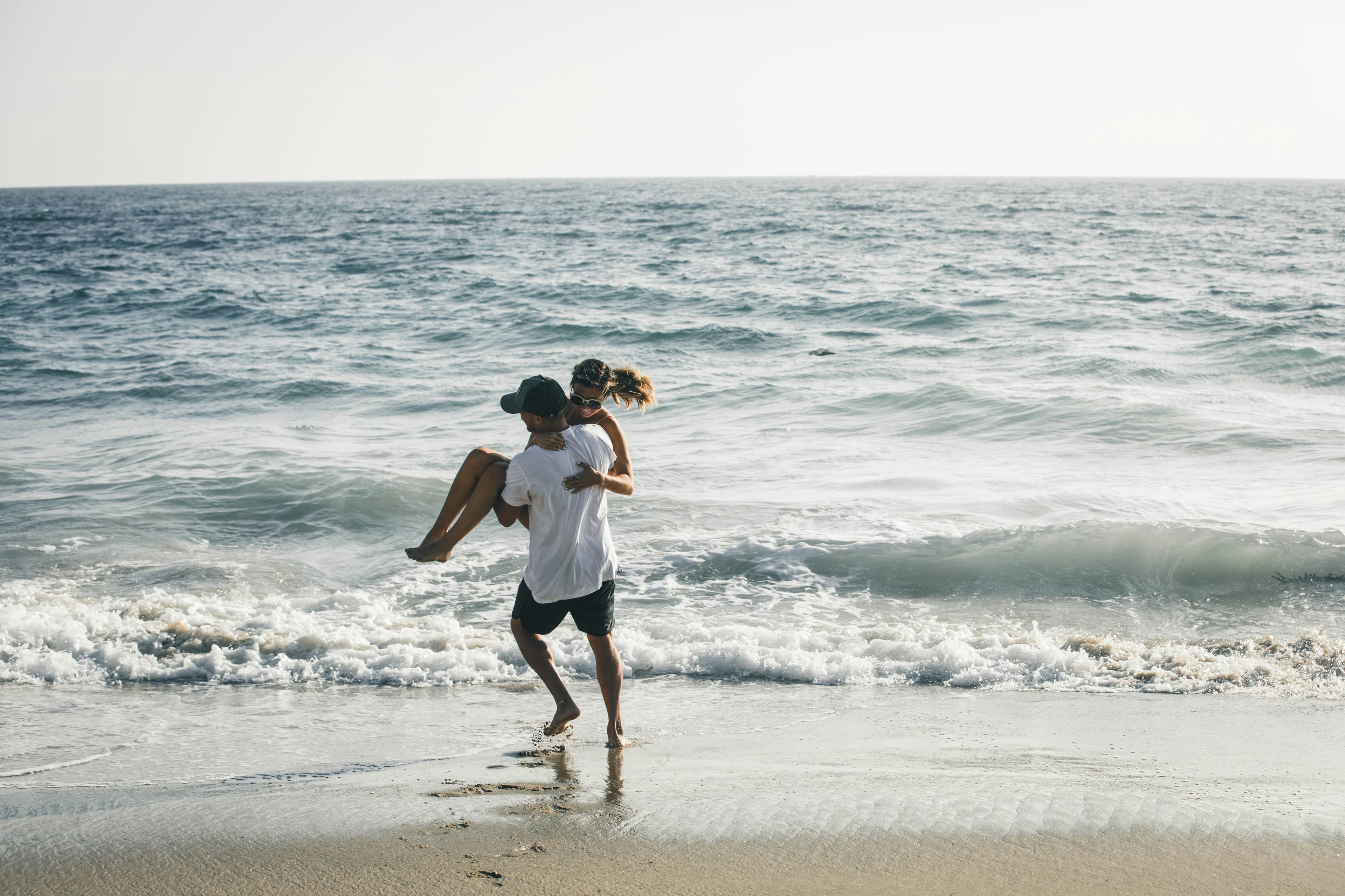 A couple plays at the beach, with one person carrying the other while walking along the shoreline as waves crash behind them.
