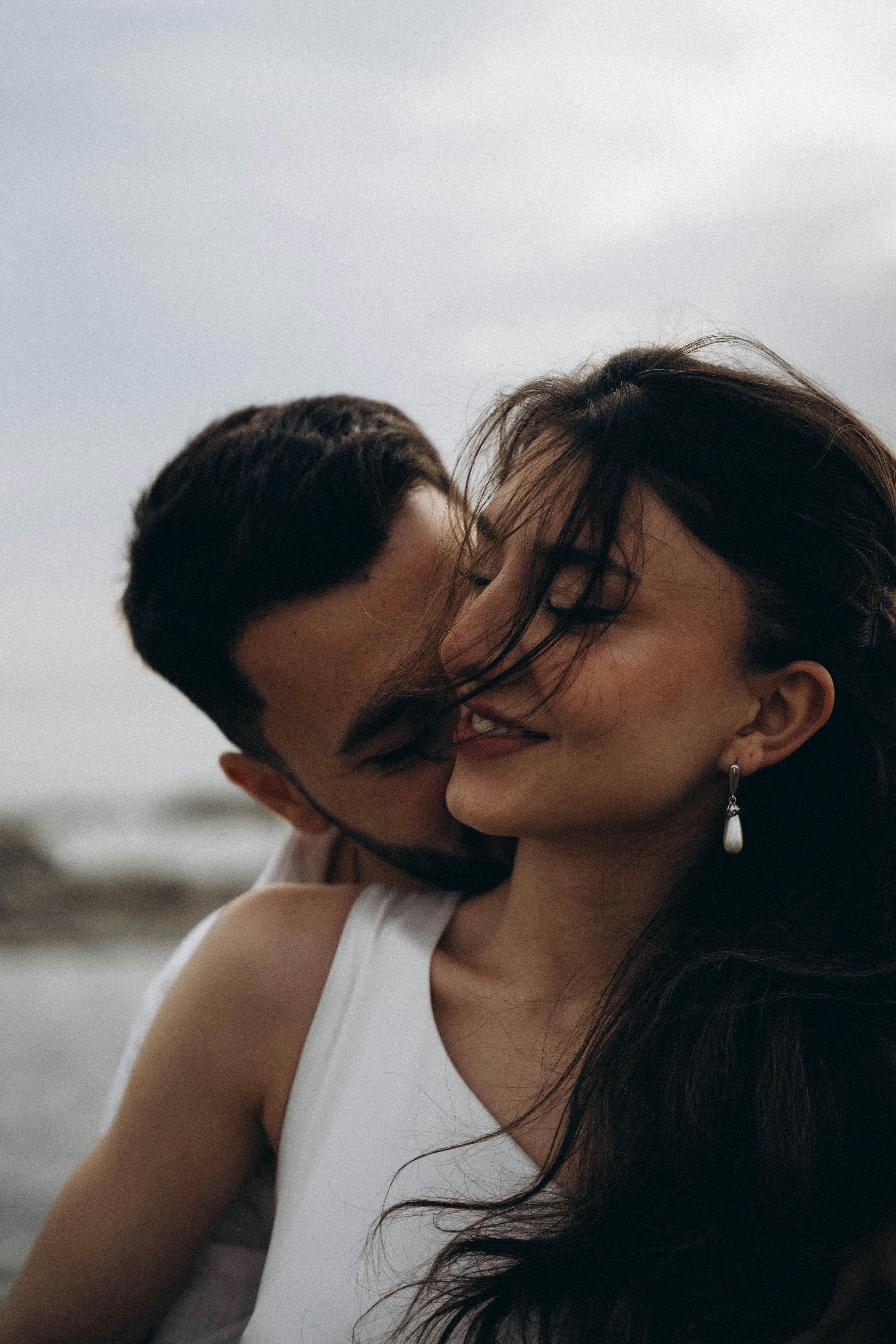 A close-up of a couple in white clothing, with wind-blown dark hair, sharing an intimate moment outdoors on an overcast day.