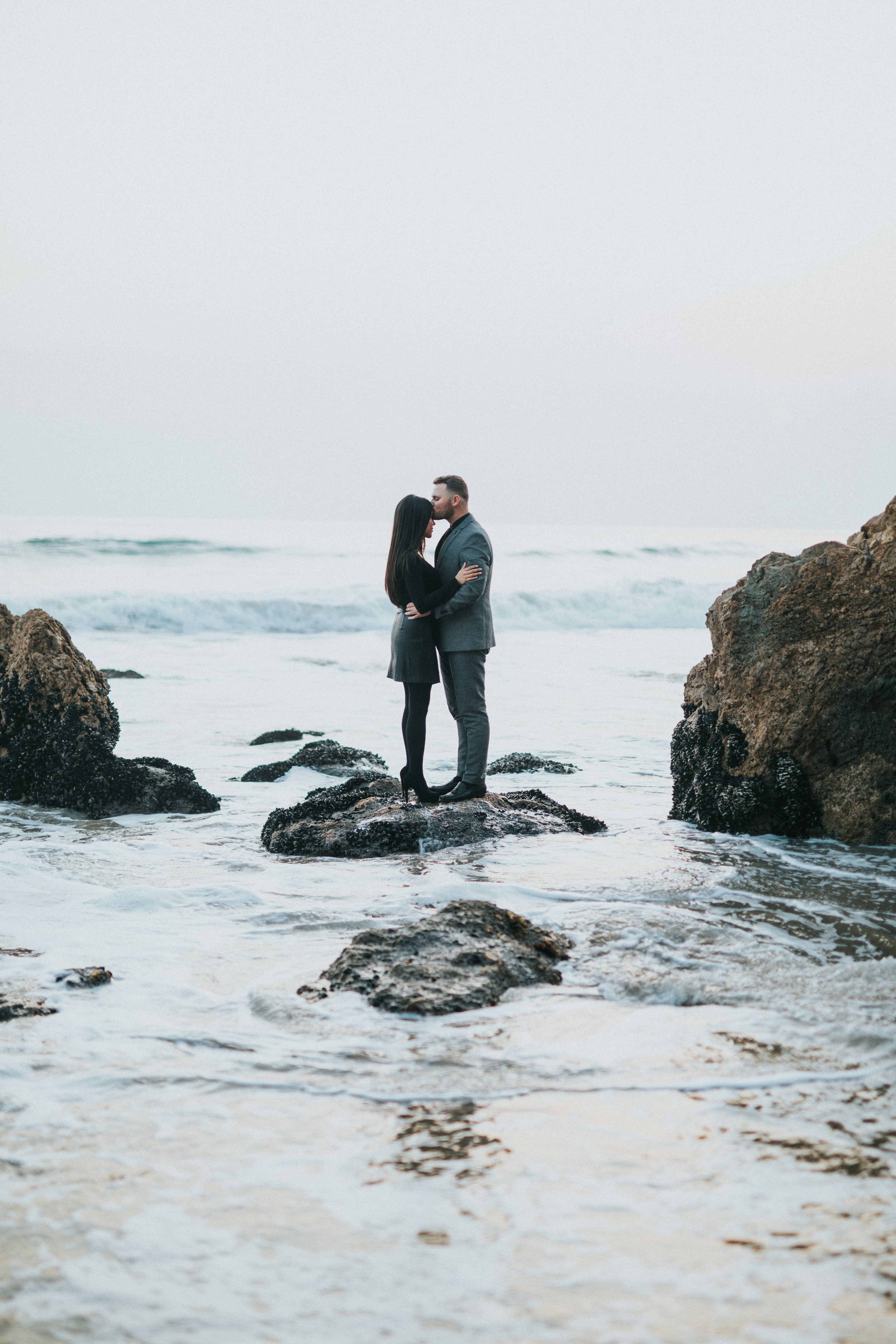 A romantic couple embraces while standing on rocky outcropping by the ocean, waves crashing around them. Moody overcast sky sets the atmosphere.