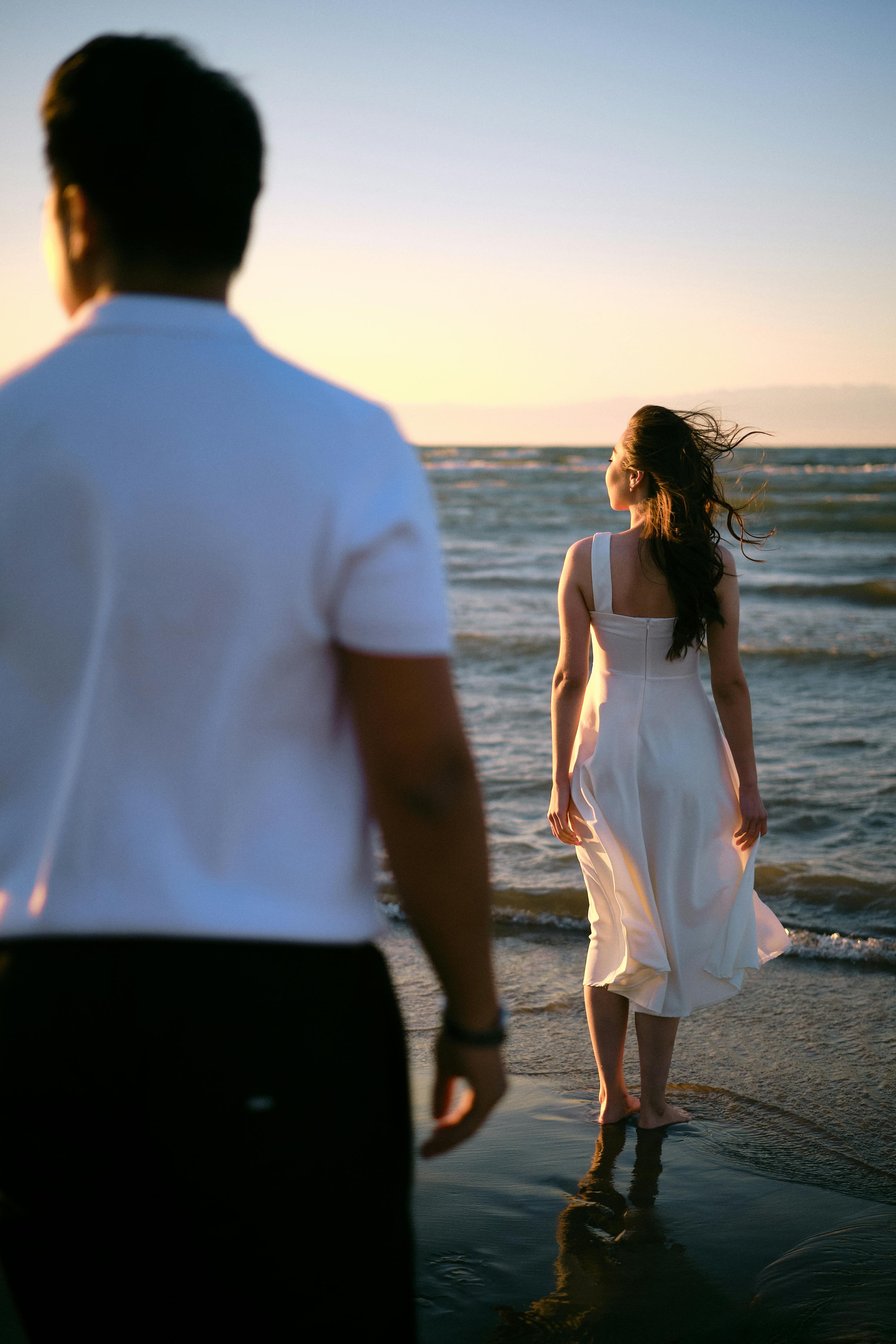 A couple on a beach at sunset - woman in white dress standing in water while man in white shirt watches from shore, her hair flowing in breeze.