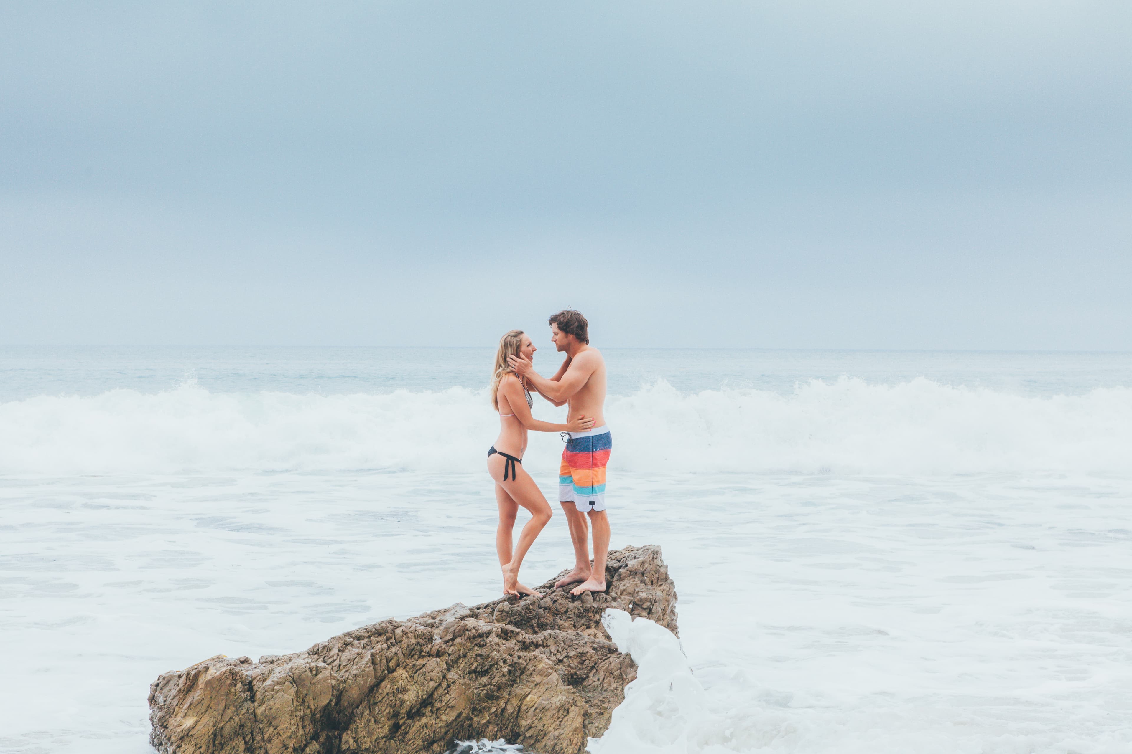 A couple in swimwear standing together on a rocky outcrop overlooking the ocean waves, sharing an intimate moment against a pale blue sky.