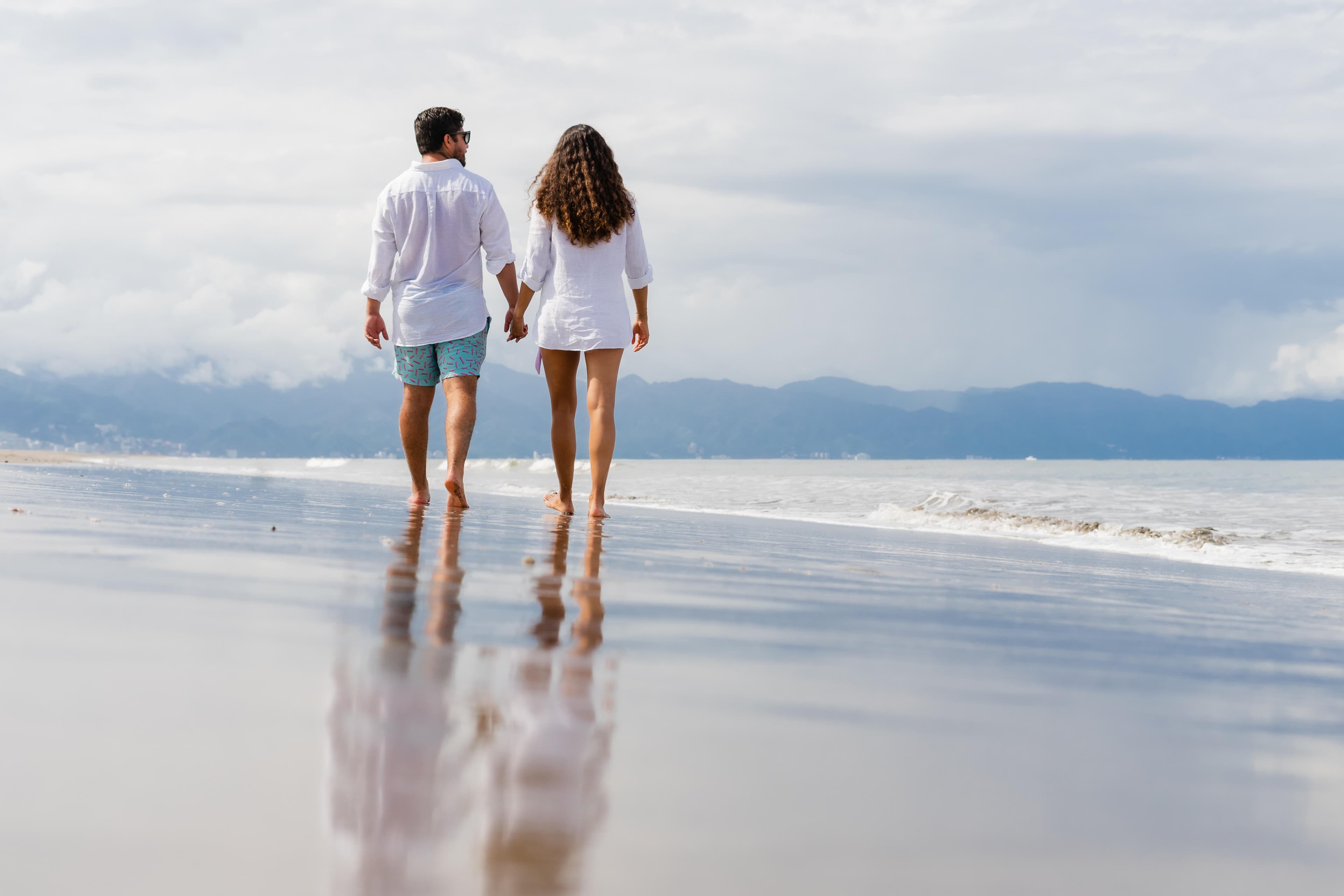 A couple in white shirts walks barefoot along a beach shoreline, their reflection visible in the wet sand. Mountains visible in the background.