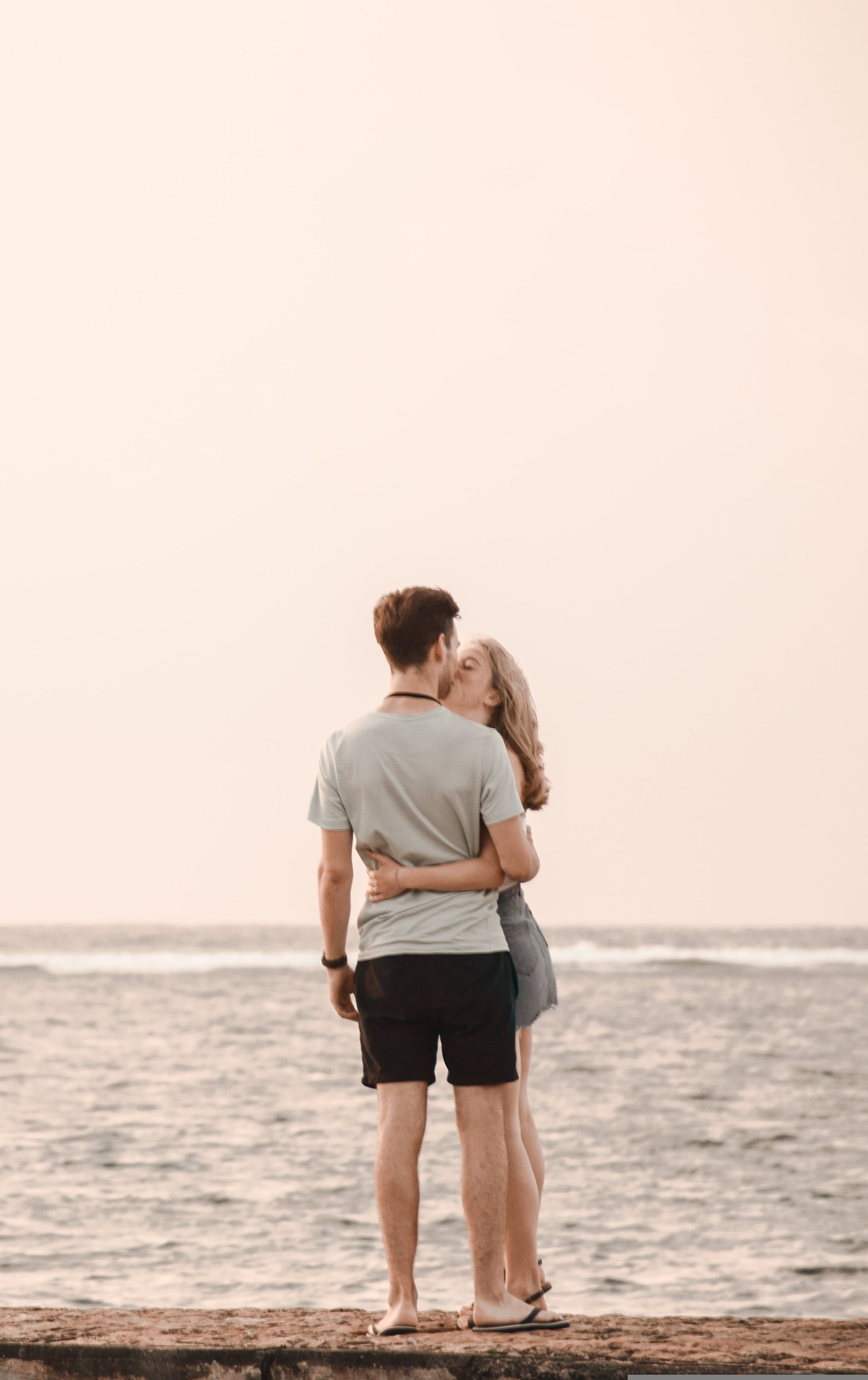 A romantic couple embracing on a beach at sunset, silhouetted against the ocean. The man wears a light t-shirt and shorts while the woman wears a casual dress.